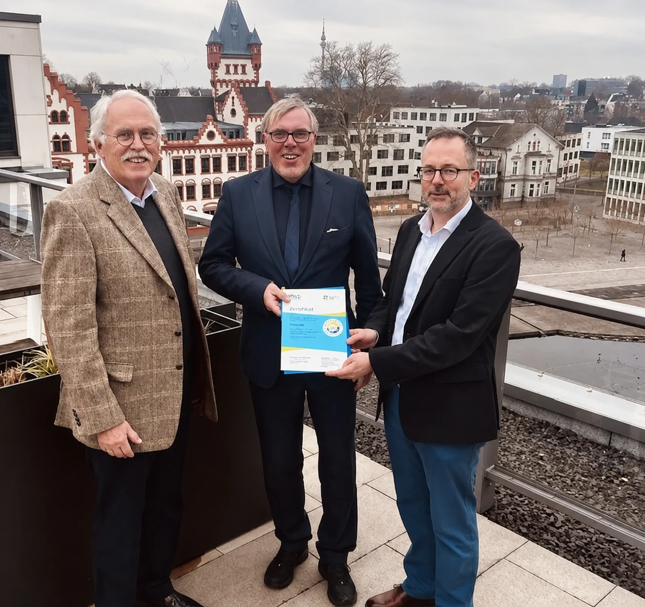 Drei Männer auf einer Dachterrasse mit Detmolder Stadtbild im Hintergrund halten das Zertifikat des Gütesiegels für Bürgerstiftungen