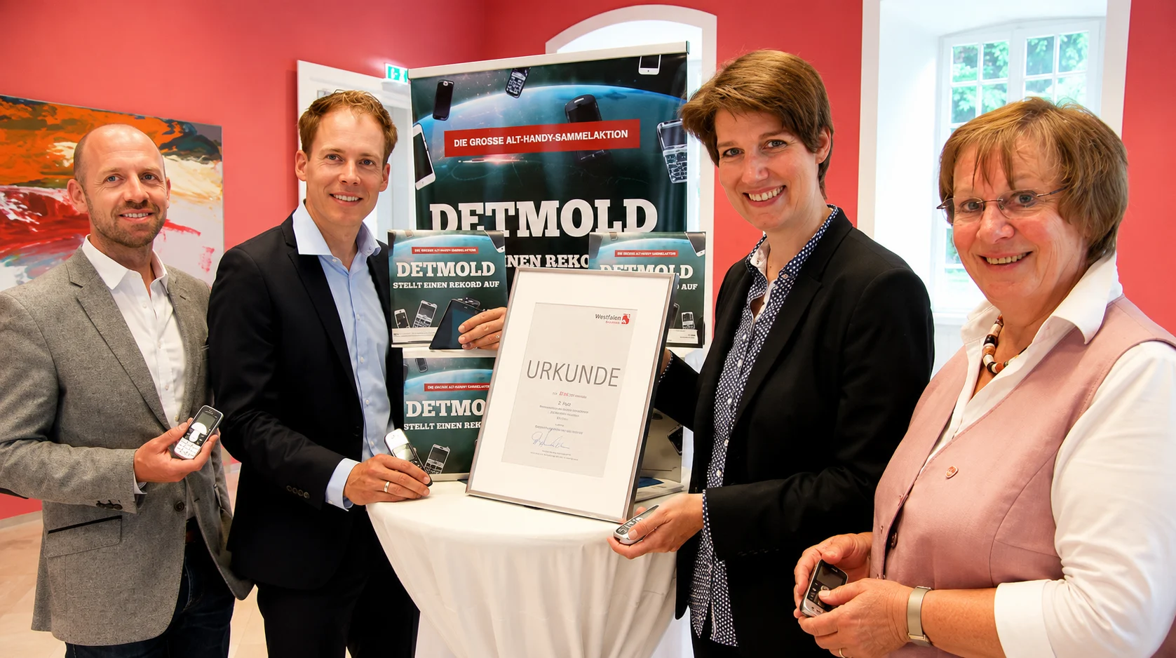Übergabe der Urkunde „Westfalen bewegt“ vor Plakaten der großen Alt-Handy-Sammelaktion in Detmold