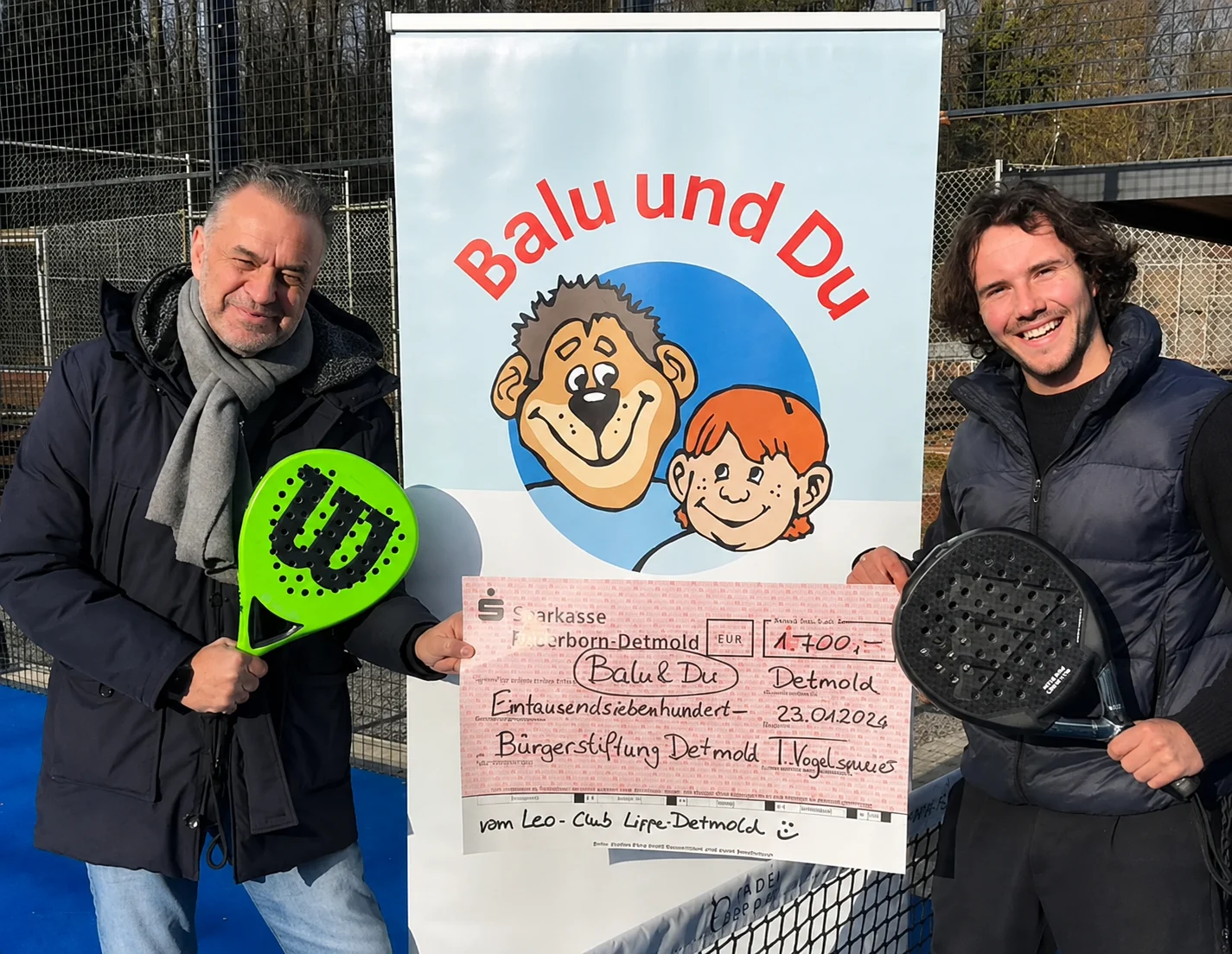 Zwei Männer mit Padel-Schlägern halten einen Spendenscheck über 1.700 Euro für das Projekt „Balu und Du“ vor dem Projektlogo