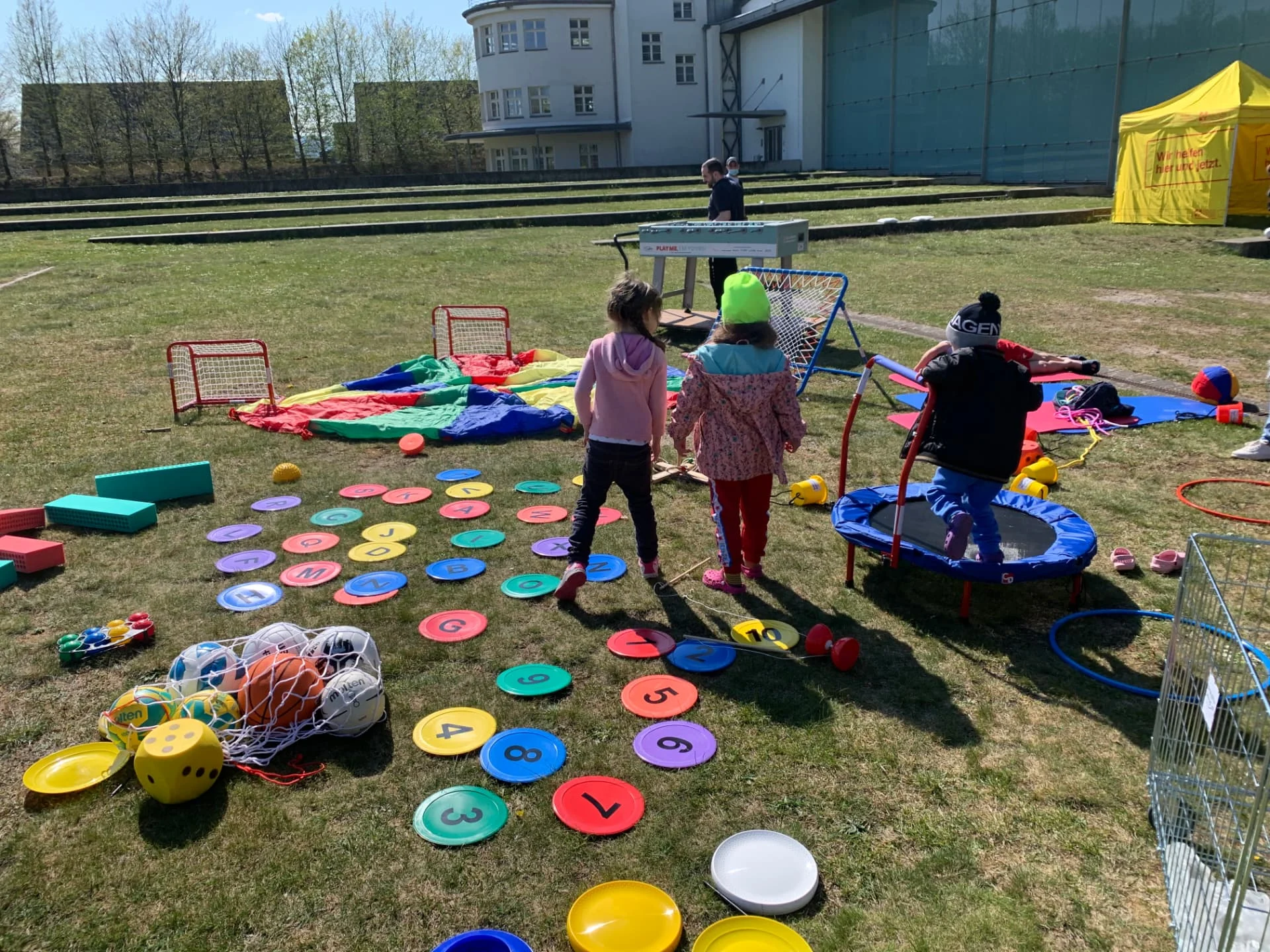 Kinder spielen auf der Wiese an der Notunterkunft Hangar 21 mit Bällen, Trampolin, Schwungtuch und bunten Bewegungsspielen