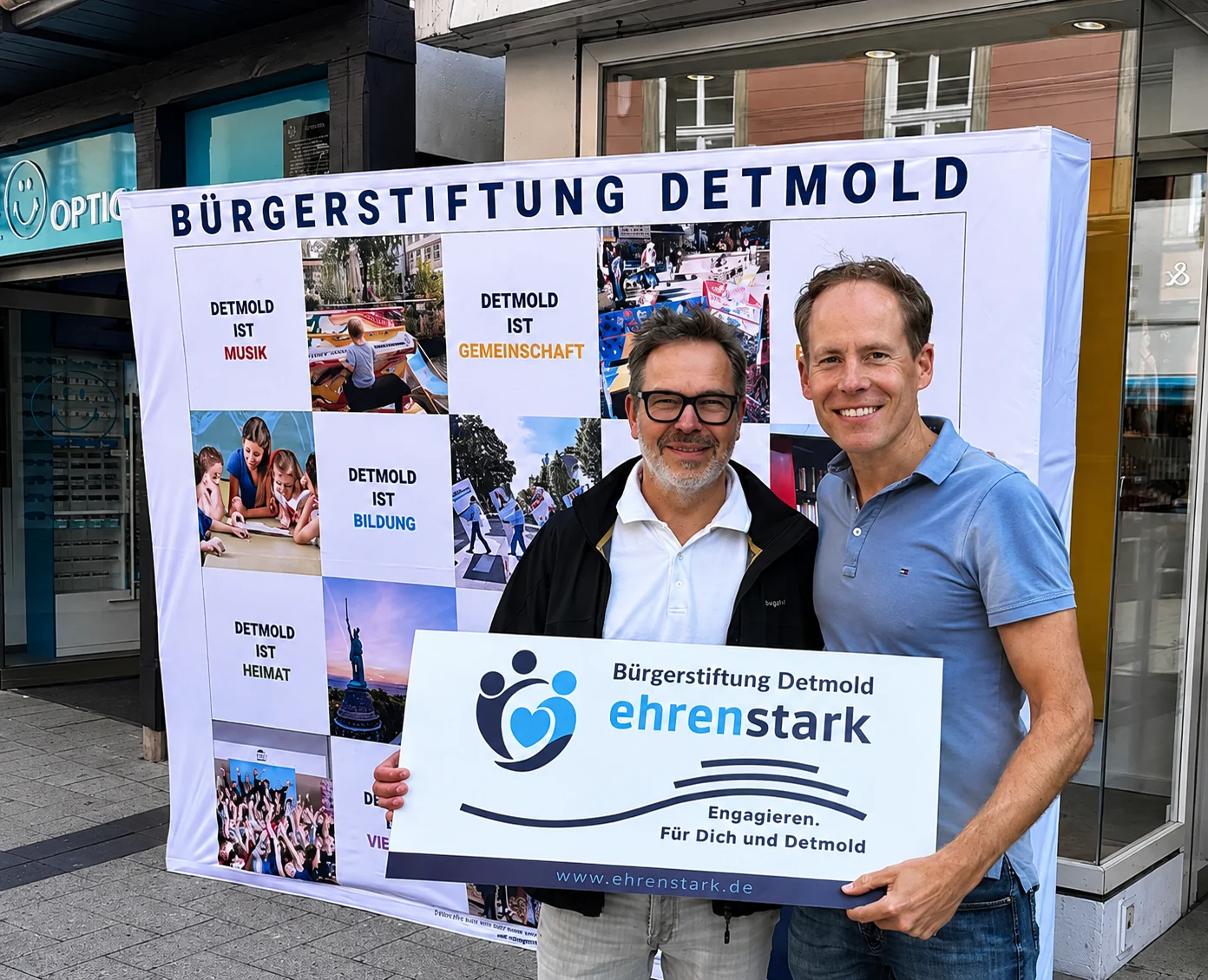 Zwei MĂ€nner halten das Schild âBĂŒrgerstiftung Detmold ehrenstarkâ vor der Fotowand der BĂŒrgerstiftung